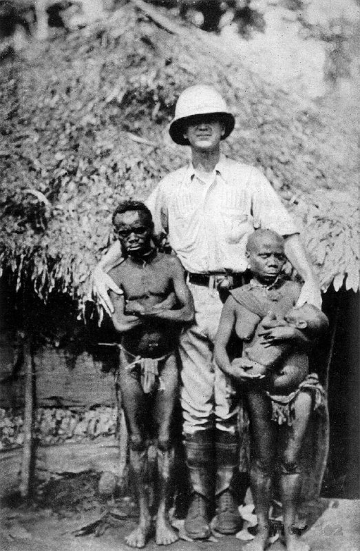1921年的这张照片显示了一户俾格米人与一名欧洲男子的身高对比，反映了他们矮小的体格特征，同时展示了当时人类学对不同族群的观察研究