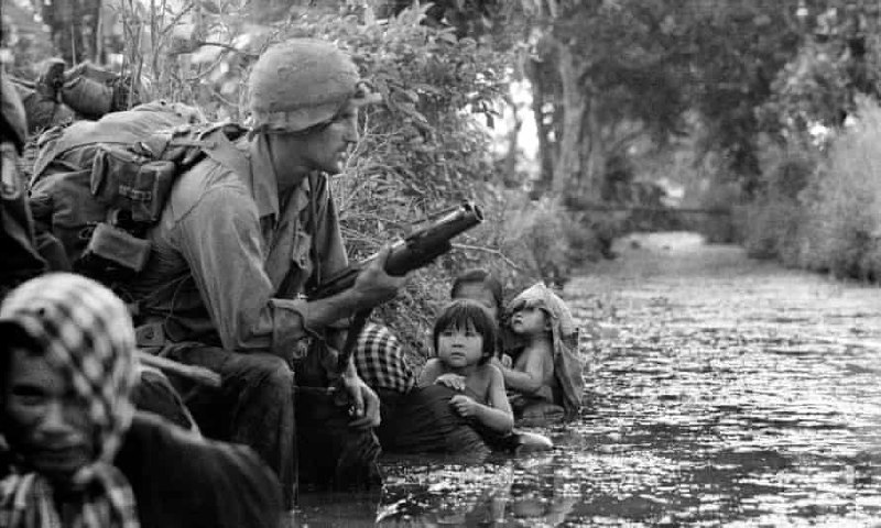 1966年，在 越南战争 期间，南越 西贡（今 胡志明市）保寨地区，一名美国伞兵手持 M79榴弹发射器 警戒，附近的孩子依偎在母亲身边，躲在运河岸边以防 越共 狙击手的袭击