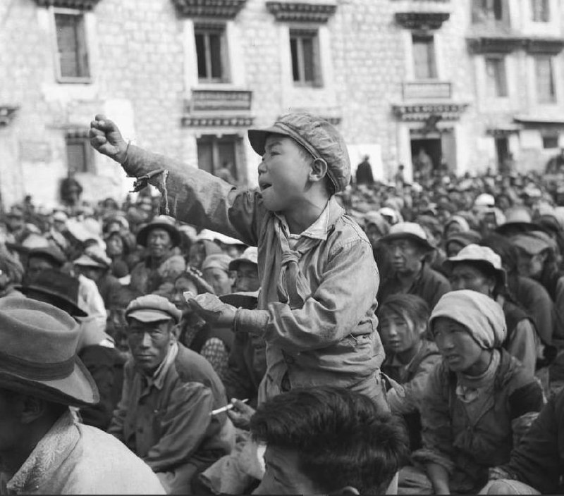 拉萨，1966或1967年，文化大革命期间举起拳头的男孩，在拉萨大昭寺教学院的集会或斗争会上，小男孩拿着一张纸片高喊口号