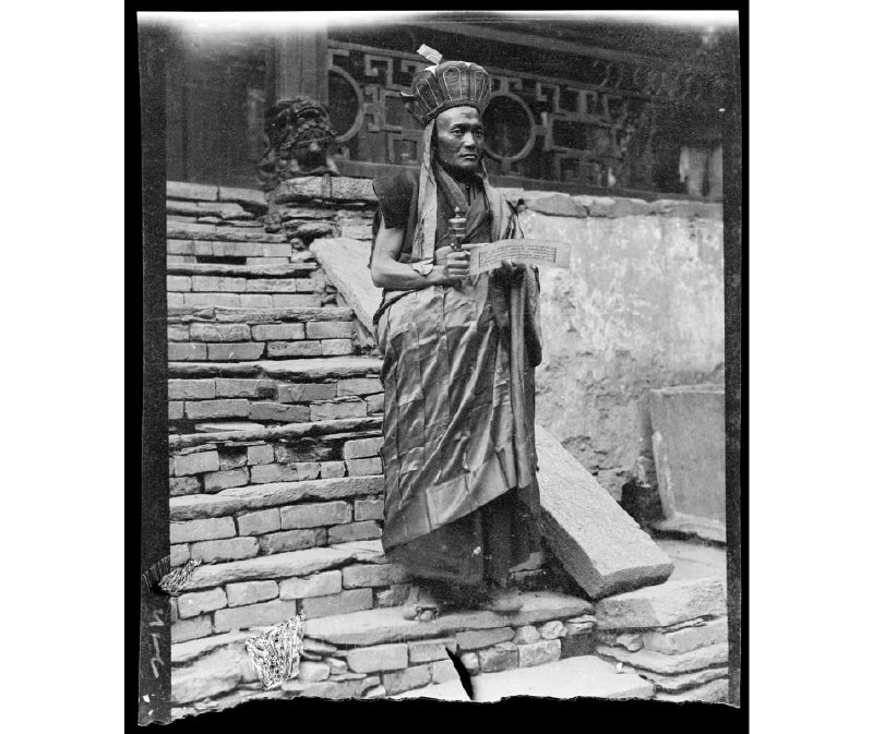 1917年四川省汶川县，绵虒镇瓦寺土司官寨，站在台阶上的一位喇嘛，头戴毗卢帽，身披袈裟，一手捧着经文，一手持着转经筒